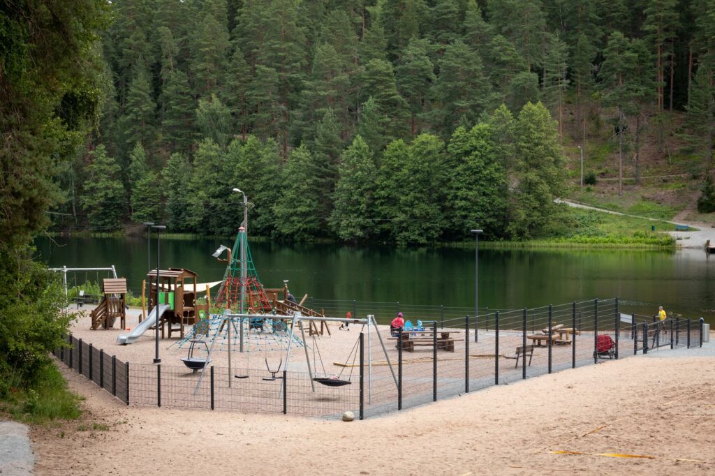 Järven rannalla sijaitseva aidattu leikkipuisto, jossa liukumäkiä, kiipeilytelineitä, keinuja sekä pöytäryhmiä.