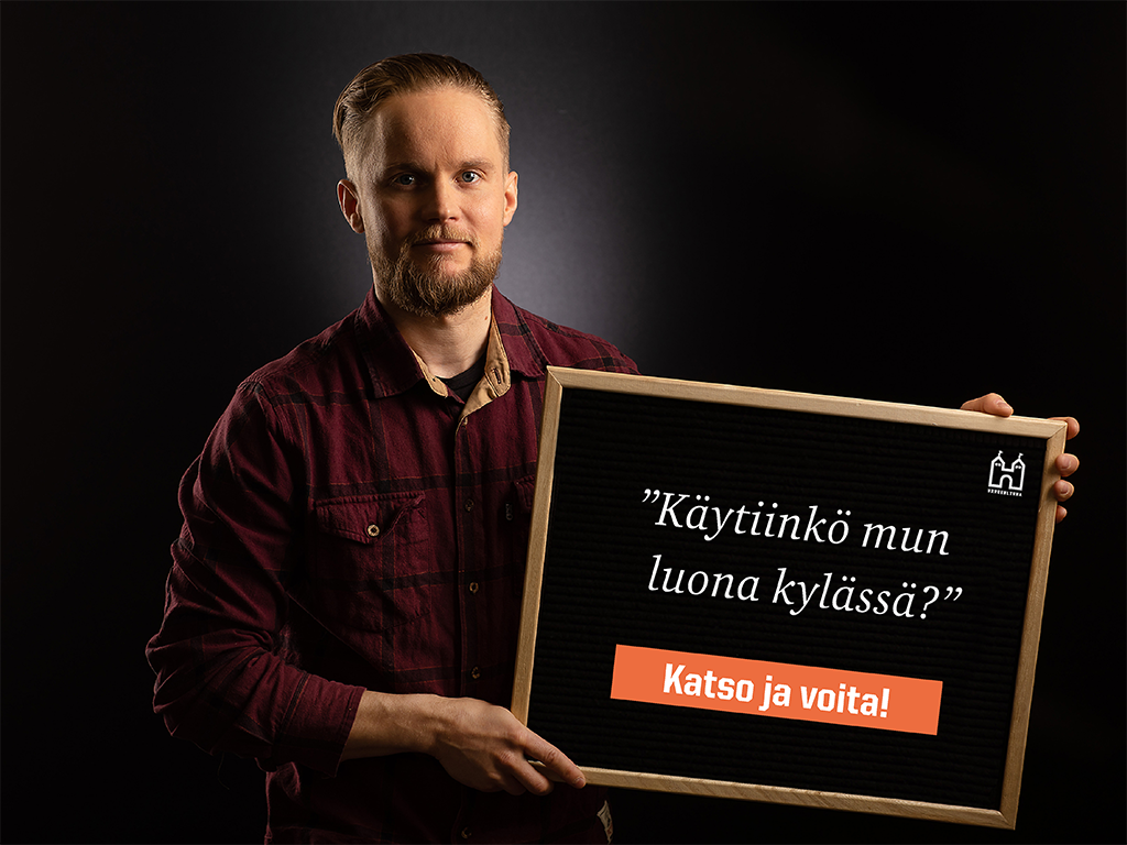 Flanellipaitainen mies pitää kädessään taulua, jossa lukee Käytiinkä mun luona kylässä? Katso ja voita!