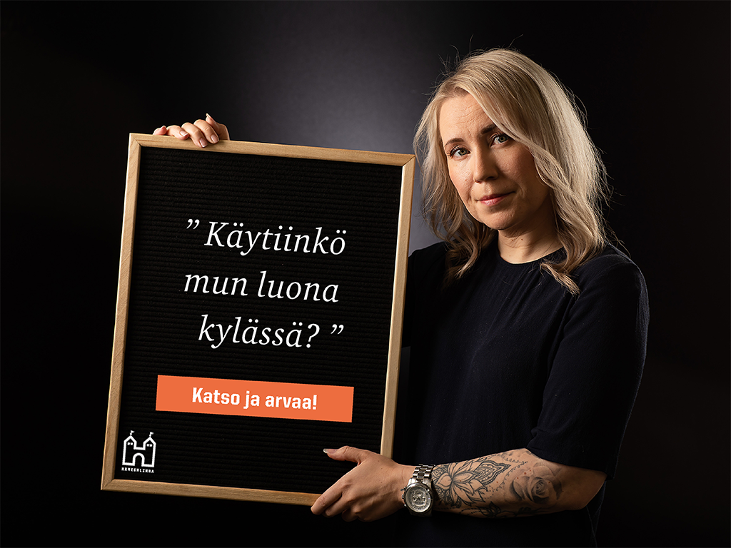 Nainen pitelee kehyksiä, joiden sisällä on Hämeenlinnan logo ja tekstit Käytiinkö mun luona kylässä? Katso ja arvaa!