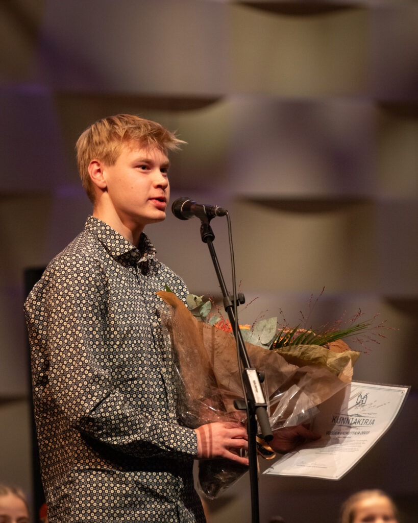 Nuori mies seisoo mikrofonin edessä ja sylissään hänellä on kukkakimppu sekä kunniakirja.