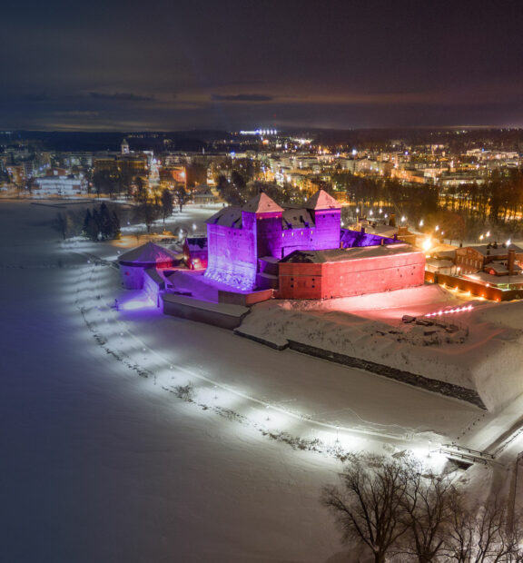 Violetiksi ja punaiseksi valaistu Hämeen linna ilmasta kuvattuna, taustalla näkyy kaupungin valot
