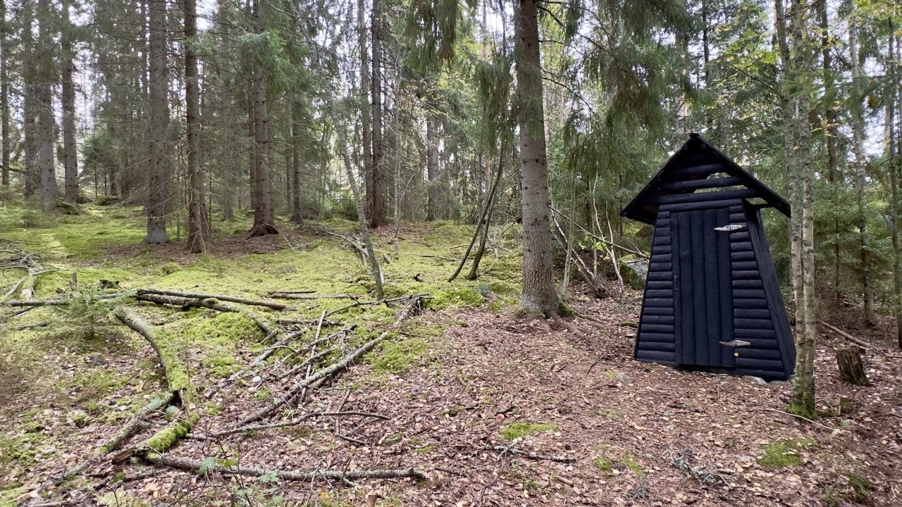 Mustaksi maalattu puinen kuivakäymälä metsässä.