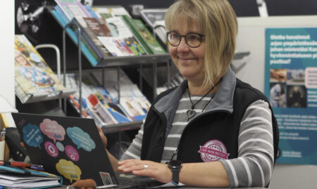 Henkilö tekee töitä tietokoneella, taustalla esitteitä.