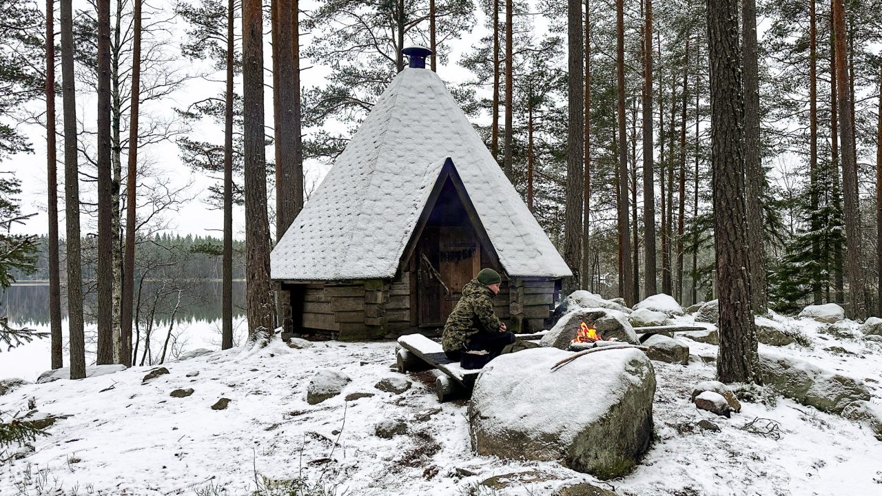 Boy sits by the fire behind a large rock, in front of the hut.