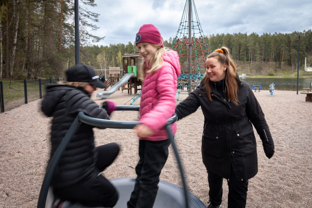 Kaksi nauravaa lasta pyörii Ahveniston leikkipaikan karusellissa, jota vieressä oleva aikuinen pyörittää.