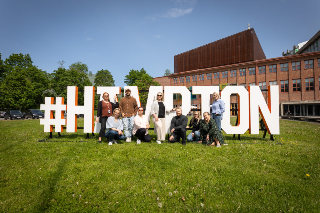 Hämeenlinnan kaupungin viestintä- ja markkinointitiimi poseeraa Verkatehtaan punatiilisen rakennuksen edessä olevien Hämpton-kirjainten juurella.