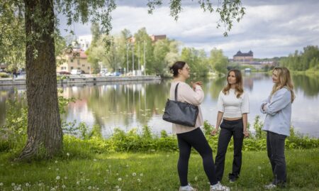 Kolme henkilöä seisoo keskustelemassa järven rannalla. Taustalla näkyy punatiilinen Hämeen linna.