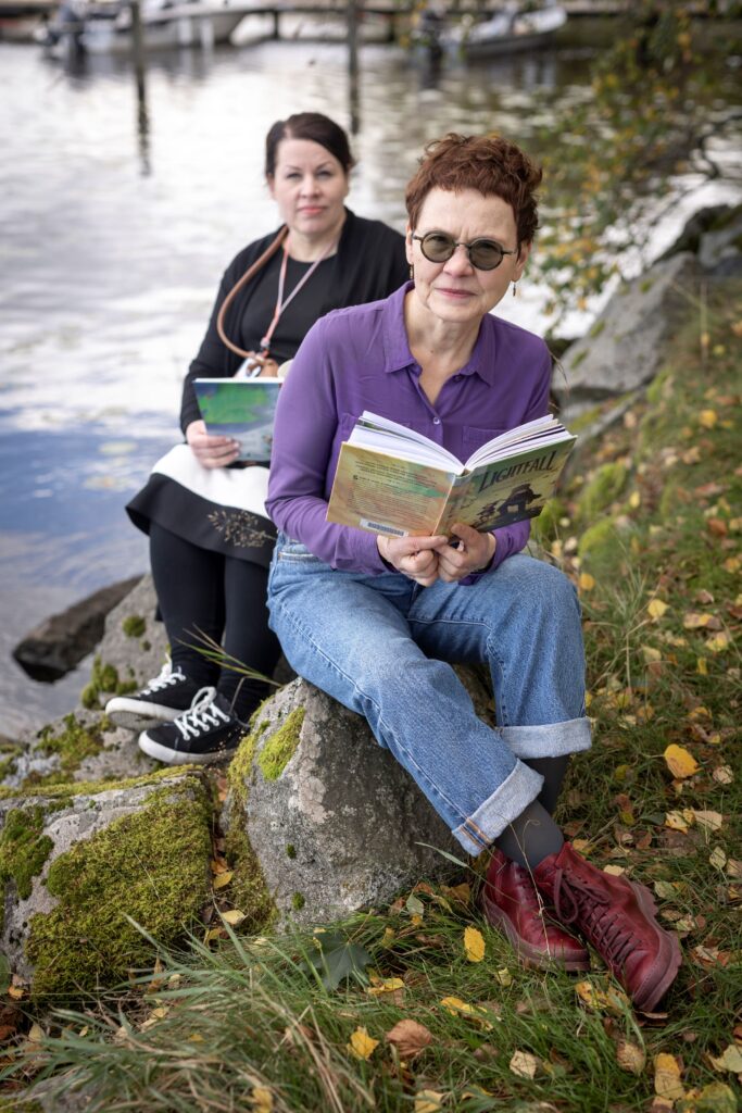 Kaksi henkilöä istuu järven rannalla lukemassa kirjoja.