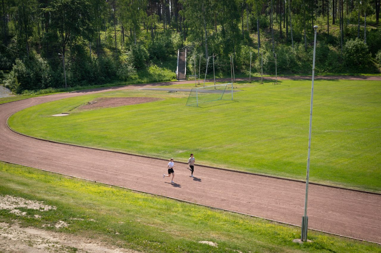 Hauhon urheilukenttä. Juoksuradalla 2 henkilöä juoksemassa. Kentän takana nousee kuntoportaat.