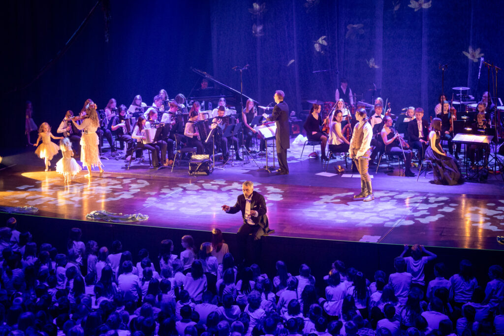 kuoronjohtaja johtaa suurta lapsikuoroa, lavalla tanssijoita, Sibelius ja orkesterit. Sininen valo.