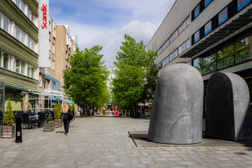 The city's pedestrian street on a sunny day. Two stone artworks and the hotel with its terraces.