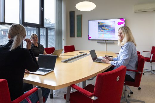 A meeting room where three people are holding a meeting in a group space. Large windows and a TV screen in the background.