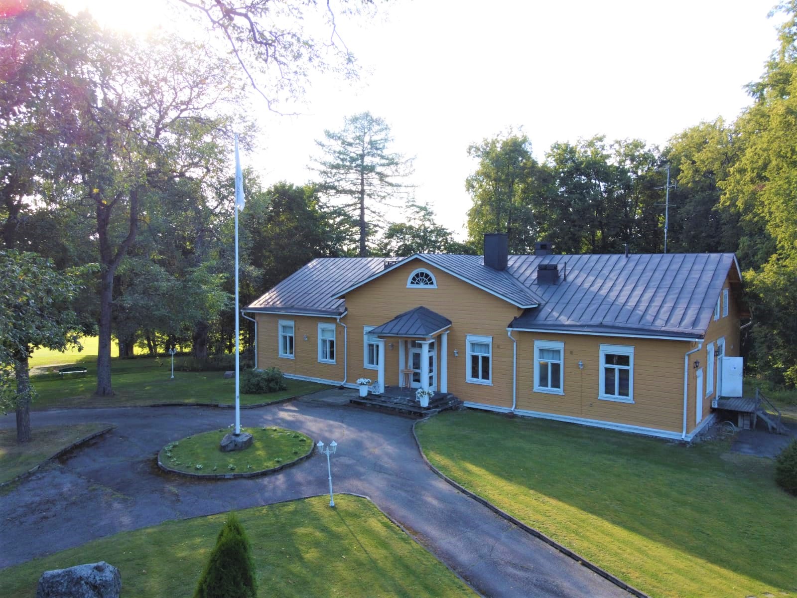 Ilmakuva keltaisesta isosta hirsisestä kartanorakennuksesta vehreässä pihapiirissä.
