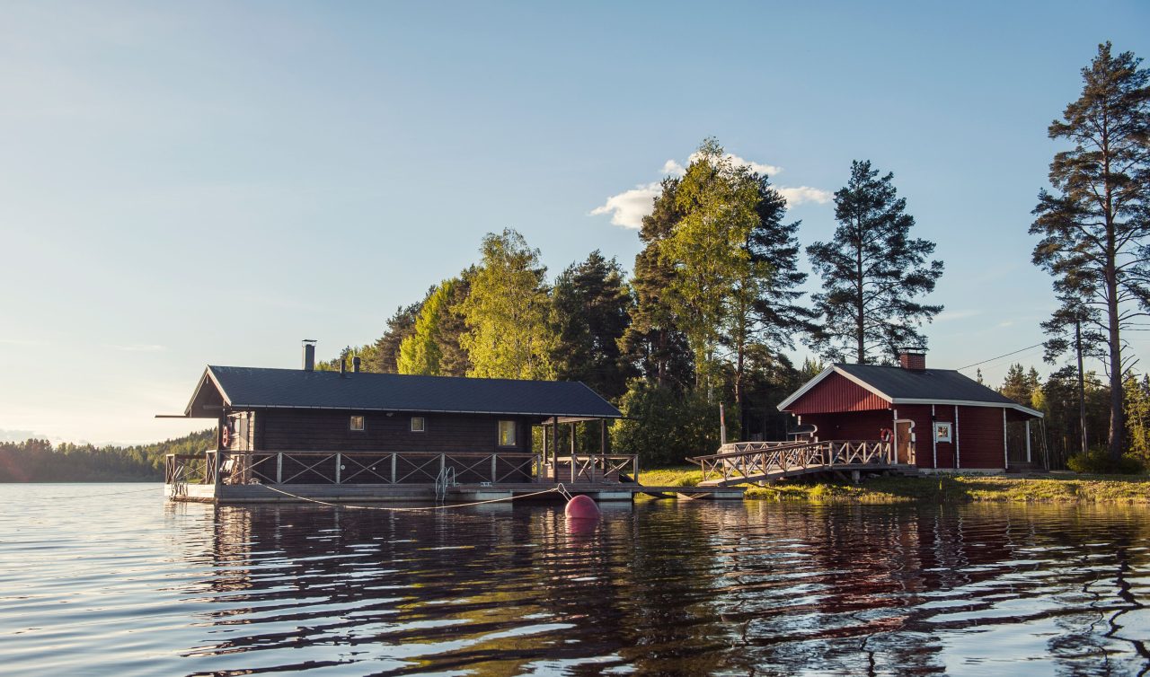 A wooden floating sauna in front of a lakeside sauna. Summer light and a lake view.
