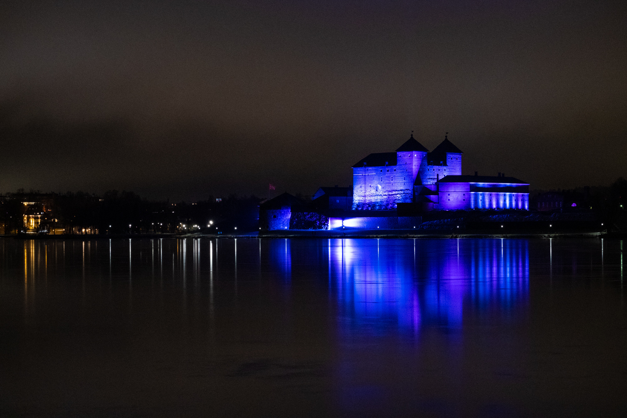 The castle illuminated in blue light at night, with the calm Vanajavesi lake in the foreground.