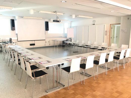 A bright meeting room with light-colored tables in a U-shape. The meeting room is ready before the arrival of clients. A whiteboard is visible in the background.