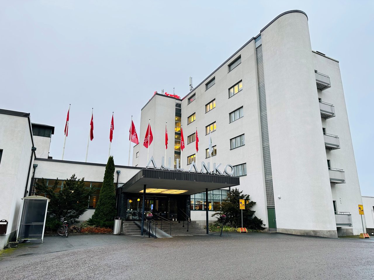 Aulangon hotellin edestä otettu kuva, jossa harmaan rakennuksen korkea oikeapuoli ja tasakattoinen sisäänkäynti. Punaiset Scandic liput keskellä rakennusta.
