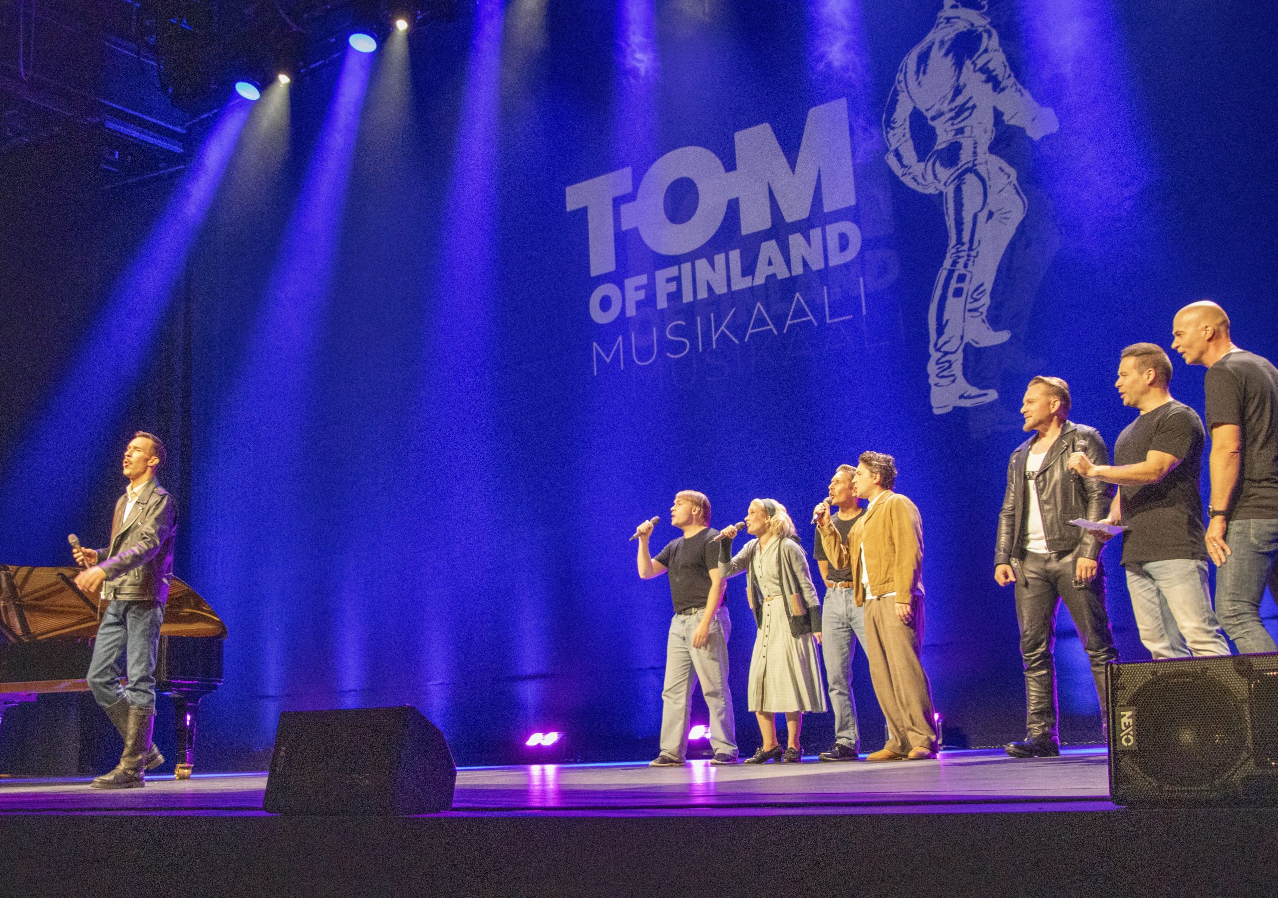 Esiintyjät lavalla Tom of Finland ‑julkistustilaisuudessa, värivalokeilat katosta ja taustalla heijastettu teksti ‘Tom of Finland’.