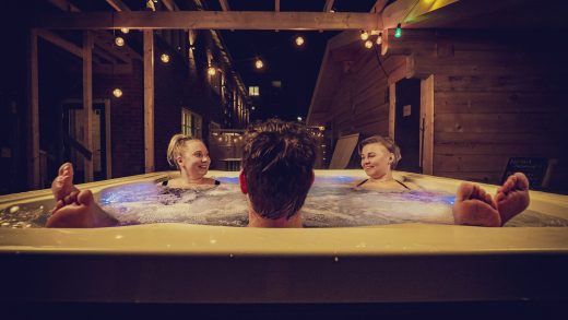 Three people in an outdoor hot tub. Hanging lights illuminate the area.
