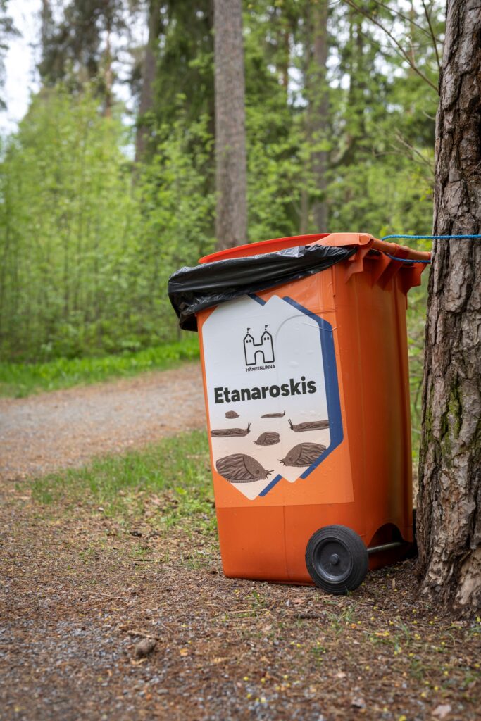Puuhun narulla kiinnitetty oranssi roskalaatikko, jonka kyljessä on Etanaroskis-tarra.
