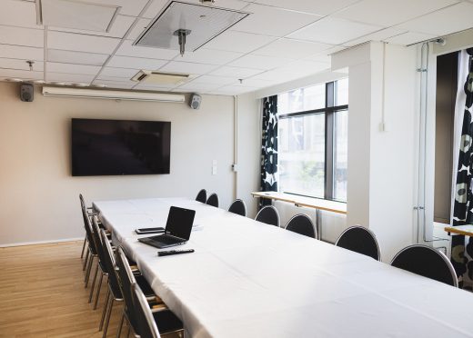 A meeting room at Hotel Emilia. A long white table with one laptop. A large screen on the back wall.