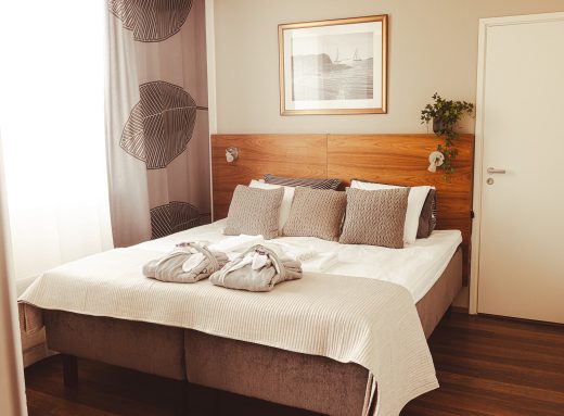 A photo of a hotel room with a made-up double bed, a painting on the back wall, and a window covered by curtains on the side.
