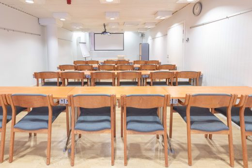 A meeting room in a classroom layout. Brown tables and chairs. A screen and flipchart on the back wall.
