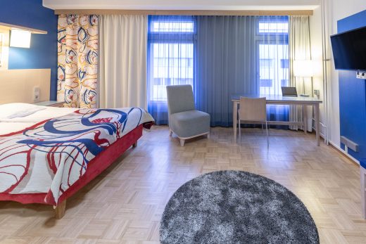 A photo of a superior room with a double bed, a colorful bedspread, and windows and an armchair in the background.