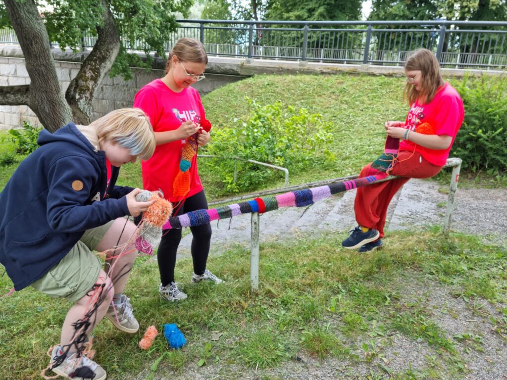 Kolme nuorta virkkaavat ja neulovat värikästä neulegraffitia